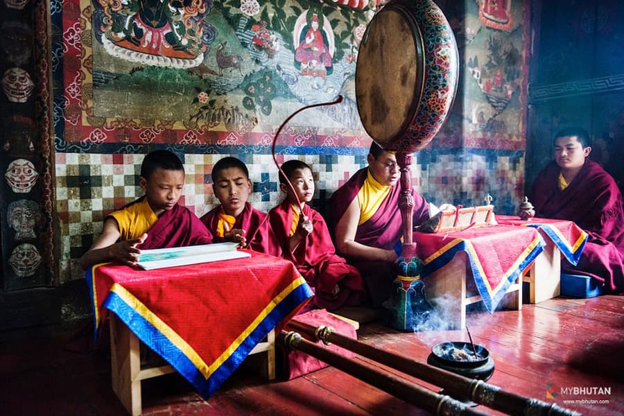 Young monks chanting from loose-leaf pecha scriptures beneath centuries-old painted murals inside the temple, one of them playing a ceremonial frame drum amid rising incense smoke.