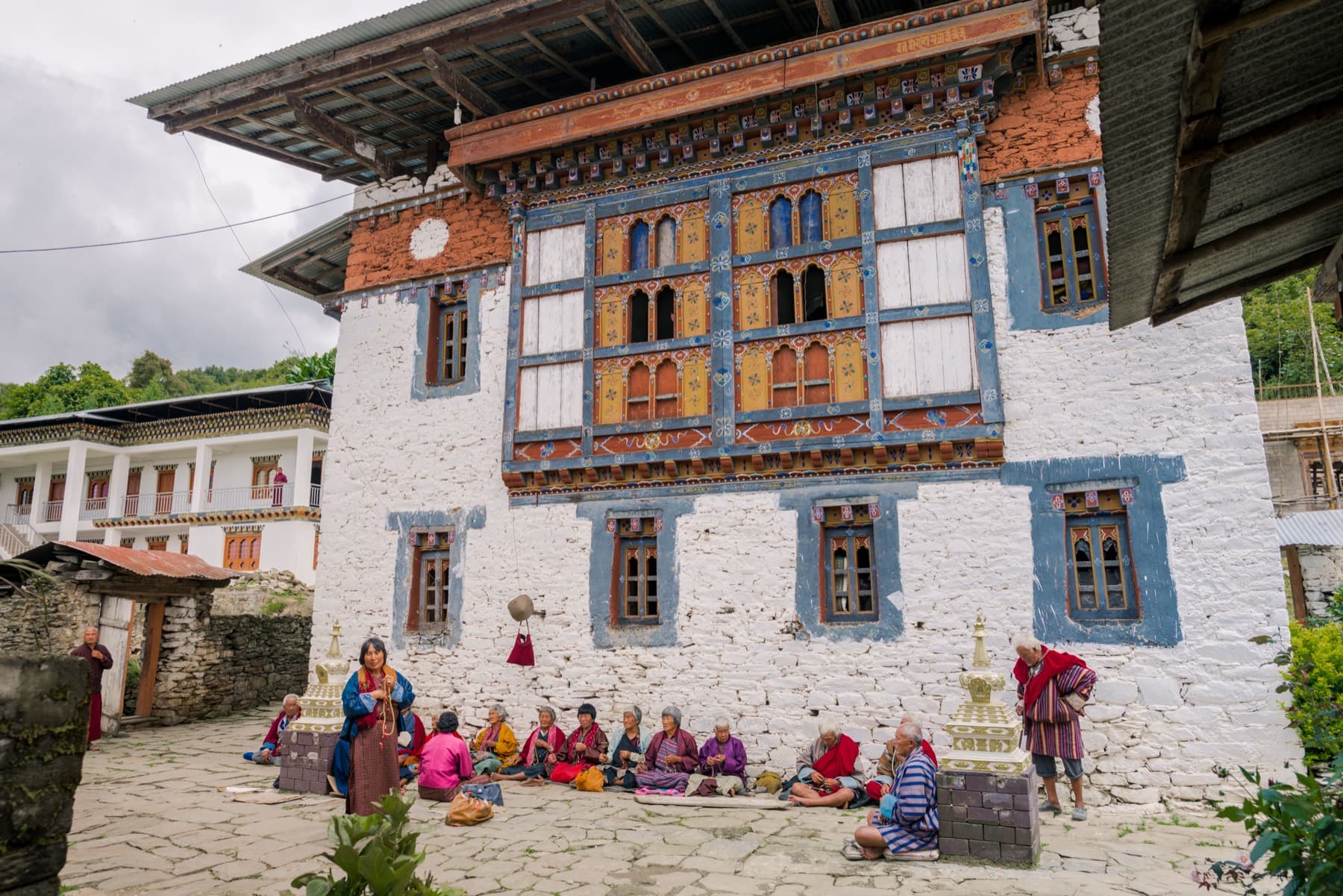 The whitewashed stone façade of Rephel Mebar Lhakhang (Sangchen Ogyen Tsuklag) with ornately painted Bhutanese window frames, small whitewashed stupas, and elderly devotees seated in the courtyard.