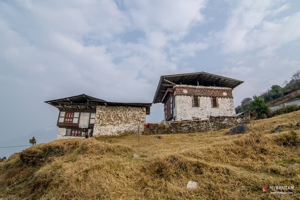 Two traditional Bhutanese stone temple buildings with painted window frames standing on a grassy hillside in the Mangde valley, Trongsa, beneath an overcast sky.